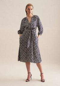 Granatowa sukienka z geometrycznym wzorem, dekoltem w serek, długimi bufkami, z dopasowaną talią i kieszeniami. Noszona z czarnymi sandałkami na obcasie.