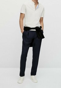 Vit kortärmad polo med ståkrage, marinblå byxor och vita sneakers. En svart tröja är knuten runt midjan. Slätt tyg, avslappnad passform.