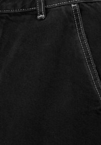 Gros plan sur un tissu en denim noir avec des surpiqûres blanches le long de la ceinture, de la passants de ceinture et du bord de la poche.