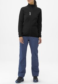 Black zip-up jacket with a high collar, paired with blue pants featuring side zippers. Model wears black hiking shoes with blue accents.