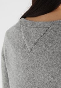 Grå stickad tröja med en triangelaccent vid nackens baksida. Mjuk textur, avslappnad passform och sömlös design. Närbild av tyget.