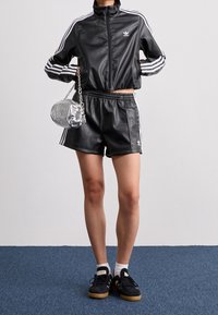 Zwarte neplederen sportjack met witte strepen en bijpassende shorts. Zilveren handtas met kettinghengsel, zwarte sneakers met een gumrubberen zool.