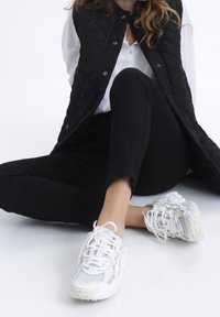 Mujer sentada en el suelo con zapatillas blancas, pantalones negros, camisa blanca y chaleco negro acolchado sobre fondo blanco.