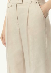 Beige Leinenhose mit Falten, seitlichen Taschen und einer subtilen Textur. Besitzt einen kleinen goldenen Akzent in der Nähe des Bundes.