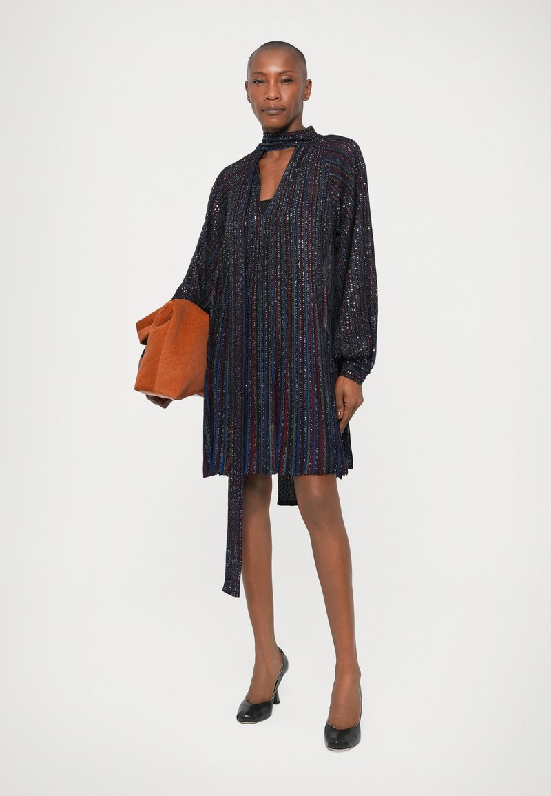 Short-sleeve, dark multicolored sequin dress with vertical stripes, a tie neck, and a cutout detail, paired with an orange clutch.