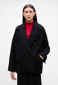 Melnā divrindas jaka ar teksturētu apdari, ar lieliem pogām un priekšējām kabatām. Valkāta uz sarkana turtlenecka.