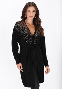 Frau in einem schwarzen langen Cardigan mit goldenen Pailletten an den Schultern, mit einem gebundenen Gürtel in der Taille, kombiniert mit schwarzen Hosen und großen Creolen.