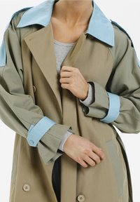 Person som knäpper en beige trenchcoat med ljusblå krage och ärmar, iförd en gråribbad långärmad tröja under.