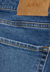 Tissu en denim avec un délavage bleu moyen, comportant des coutures orange, une poche arrière et une étiquette en cuir brun clair estampillée "JJXX."