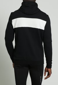 Felpa nera con una striscia orizzontale bianca lungo la parte superiore della schiena. Presenta un cappuccio con lacci e polsini e orlo a coste. Tessuto dalla texture liscia.