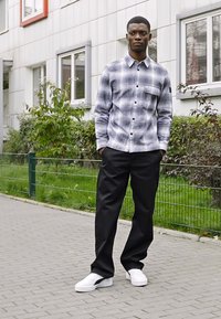 Szürke és fehér kockás ing gombokkal és zsebbel, fekete nadrággal és fehér sportcipővel párosítva. Kültéren, zöld növényzet közelében áll.
