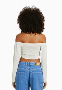 Donna con capelli ricci che indossa un crop top bianco a spalle scoperte e jeans blu a vita alta, in piedi di spalle su uno sfondo bianco.