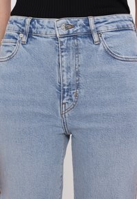 Jean en denim bleu clair à taille haute, comportant cinq poches, une fermeture à bouton en métal et des coutures visibles en orange contrastant.