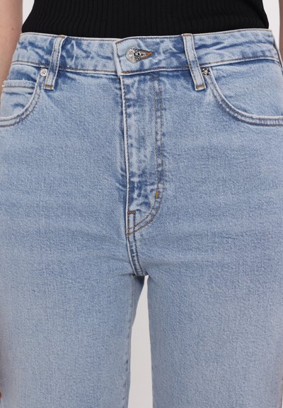 Valkoinen denim-farkut korkealla vyötäröllä, jossa on viisi taskua, metallinappi suljettuna ja näkyvää ompelua kontrastivärisessä oranssissa.