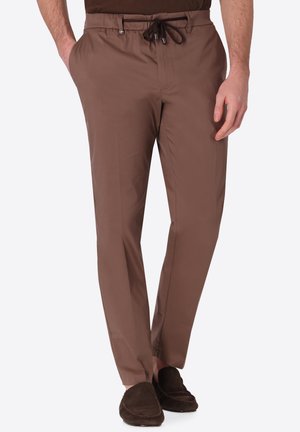 Uomo che indossa pantaloni marroni slim fit con vita con coulisse e scarpe slip-on marrone scuro, in piedi con una mano in tasca.