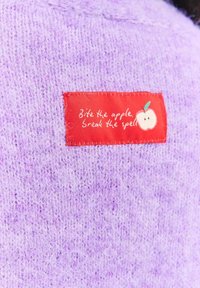 Fioletowa dzianina z naszywką w kolorze czerwonym, na której znajduje się biały graficzny obraz jabłka oraz tekst „Ugryź jabłko, przełam zaklęcie.”