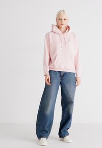 Light pink hooded sweatshirt with front pocket, paired with oversized blue jeans. Model wearing white sneakers. Simple, casual design.