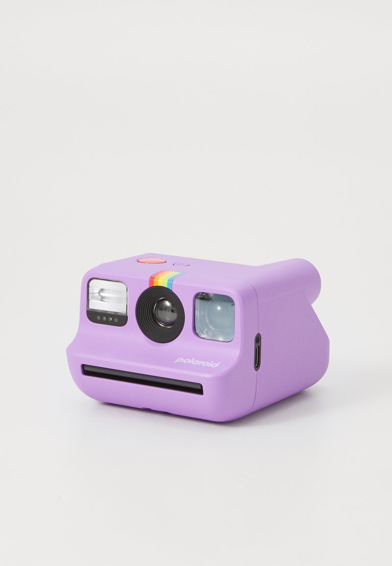 Fialový instantní fotoaparát Polaroid s duhovým pruhem, objektivem, bleskem, hledáčkem a slotem na fotografie na bílém pozadí.