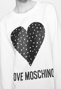 Bílá mikina s velkým černým srdcem jako grafikou, malými bílými srdíčky a textem "LOVE MOSCHINO" ve výrazných černých písmenech pod ním.