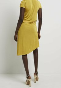 Vestido amarillo de manga corta con una silueta drapeada y dobladillo asimétrico. Presenta una textura suave y un acento metálico a la altura de la cintura.