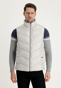 Chaleco acolchado gris claro con cuello alto y patrón en forma de chevron, que cuenta con una cremallera frontal, bolsillos laterales y una textura suave y brillante.