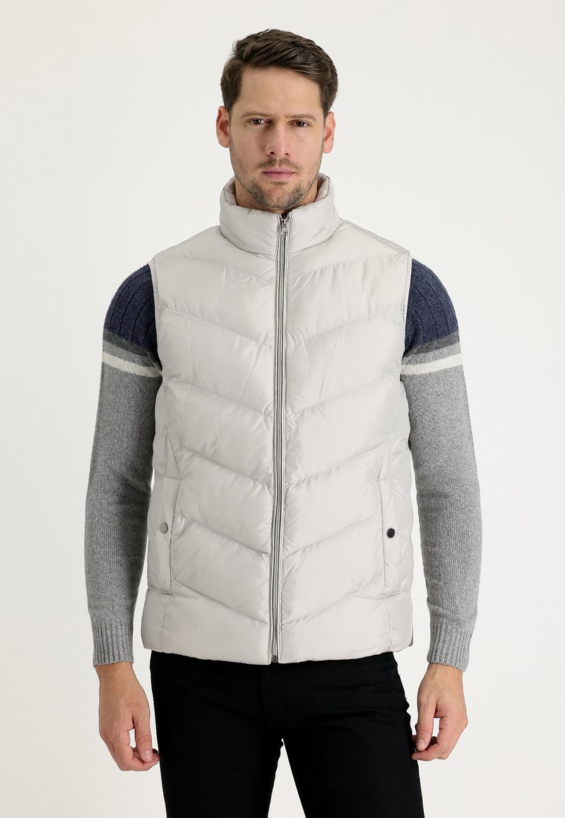 Chaleco acolchado gris claro con cuello alto y patrón en forma de chevron, que cuenta con una cremallera frontal, bolsillos laterales y una textura suave y brillante.