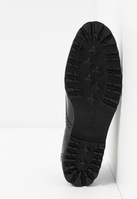Semelle de chaussure noire avec des motifs de traction prononcés. Présente une marque sur le côté et une surface texturée pour l'adhérence. Matériau en caoutchouc durable.