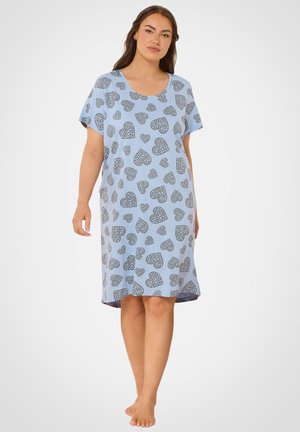 Frau trägt ein locker sitzendes, hellblaues knielanges Kleid mit grauen Herzmustern, steht barfuß vor weißem Hintergrund.