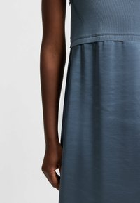 Bezramínková šaty v měkké modré tkanině s přiléhavým vrškem a splývavou sukní. Hladký povrch s kontrastní pletenou detailní zdobení na korpusu.