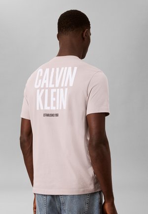 Hombre que lleva una camiseta rosa claro de Calvin Klein con un gran logo blanco y texto "Established 1968" en la espalda, combinado con vaqueros azules.