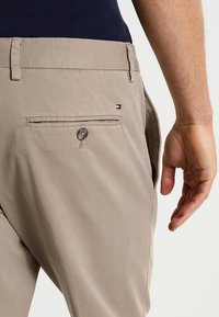 Beige broek met een achterzak voorzien van een zilveren knop en een klein logo patch. De stof lijkt glad en lichtgewicht.