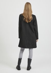 Cappotto lungo nero con taglio dritto, realizzato in un materiale morbido. Presenta maniche lunghe e lunghezza fino a metà coscia, abbinato a pantaloni slim grigi e stivali neri.