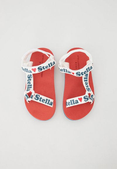 Sandalias planas rojas con tiras blancas que presentan el texto "Stella" y un detalle de corazón. La suela tiene un acento azul. Diseño de plantilla texturizada.