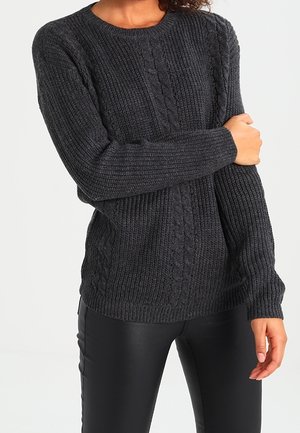 Femme portant un pull en maille torsadée gris foncé et un pantalon en cuir noir, tenant un bras croisé devant son corps.