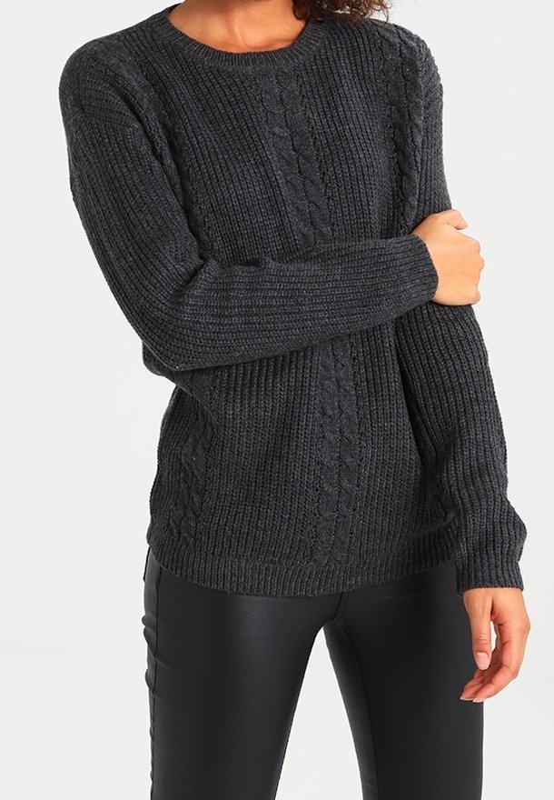 Femme portant un pull en maille torsadée gris foncé et un pantalon en cuir noir, tenant un bras croisé devant son corps.
