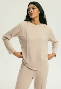 Beżowa bluzka z długim rękawem i okrągłym dekoltem, wykonana z teksturowanej tkaniny, z pionowymi przeszyciami z przodu. Stylizowana z dopasowanymi spodniami.
