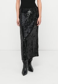 Jupe maxi noire à sequins avec une taille cintrée, présentant une texture brillante et une silhouette droite, associée à des bottines noires montant jusqu'à la cheville.