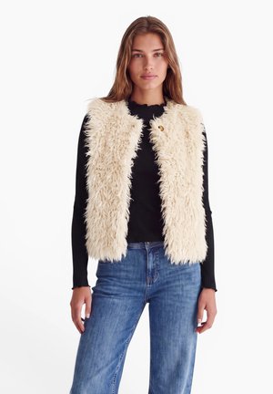 Femme portant une veste crème duveteuse sur une chemise noire à manches longues et un jean bleu, debout sur un fond blanc.