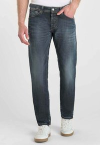 Jeans de mezclilla azul oscuro con desgaste y arrugas, corte recto, diseño de cinco bolsillos, botón de metal y detalles de costura. Combinados con zapatillas blancas.