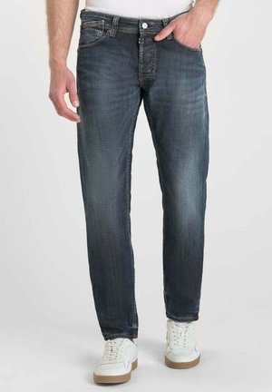 Jean en denim bleu foncé avec décoloration et effacement, coupe droite, design à cinq poches, bouton en métal et accents cousus. Portés avec des baskets blanches.
