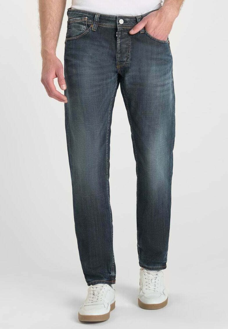 Jeans de mezclilla azul oscuro con desgaste y arrugas, corte recto, diseño de cinco bolsillos, botón de metal y detalles de costura. Combinados con zapatillas blancas.