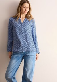 Blå mønstret langærmet bluse med V-udskæring, parret med lyseblå vide jeans, vist mod en neutral baggrund.