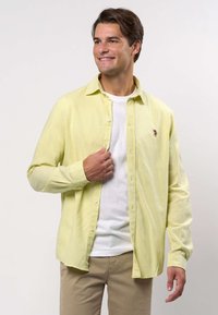 Lächelnder Mann, der ein hellgelbes Hemd über einem weißen T-Shirt und beige Hosen trägt und das Hemd mit seiner linken Hand richtet.