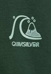Grønn stoff med en lett, stilisert bølge og vulkanlogo med bobler over, ledsaget av teksten "QUIKSILVER" under.