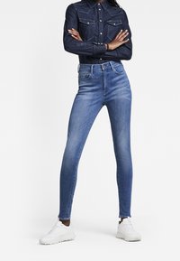 Jeans de denim ajustados en azul medio con una cintura alta y ligero despejado. Combinados con una camisa de denim oscura y zapatillas blancas.