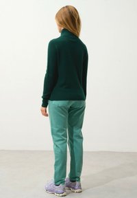 Groene gebreide coltrui, slim-fit lichtgroene broek en pastel paarse sneakers. Achtergrond is effen wit met een grijze vloer.