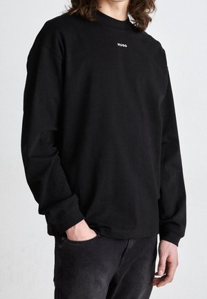 Personne portant un t-shirt noir à manches longues avec le texte "HUGO" sur la poitrine et un pantalon noir, une main dans la poche, sur un fond uni.