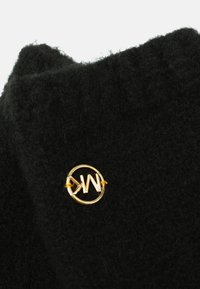 Czarna wełniana tkanina o miękkiej fakturze, z okrągłym logo z metalu w kolorze złotym, przymocowanym małą pętelką.