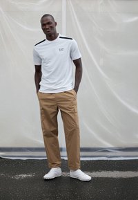 Baltas īsroku krekls ar melnām detaļām, brūnas krāsas darba bikses un baltas sporta kurpes. Gluda auduma tekstūra un brīvs piegriezums, attēlots neitrālā fonā.