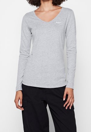 Femme portant un t-shirt gris clair à manches longues et col en V, ainsi qu'un pantalon noir avec des poches cargo, debout devant un fond uni.